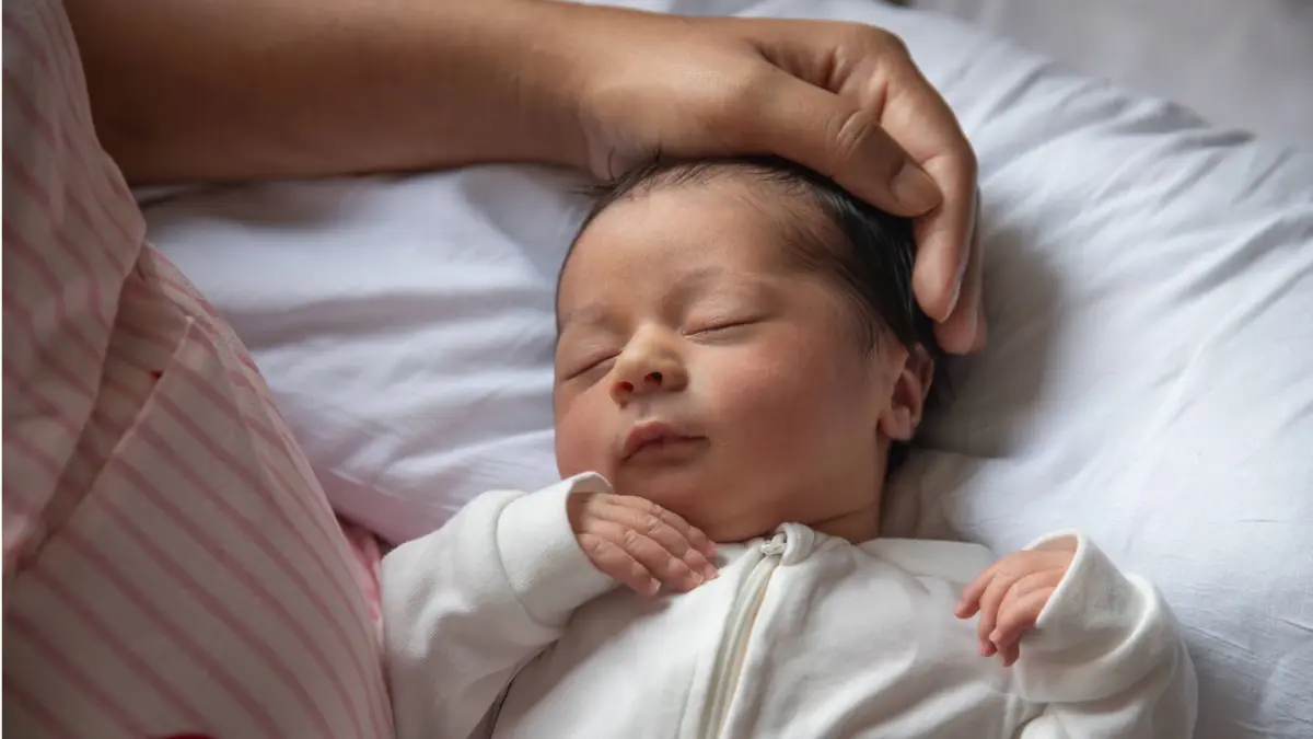 A gentle hand caresses the head of a sleeping newborn baby, dressed in a white onesie on soft bedding. Model Released TeoTarras_ID17101_521237_015 Copyright: xTeoxTarrasx RECORD DATE NOT STATED