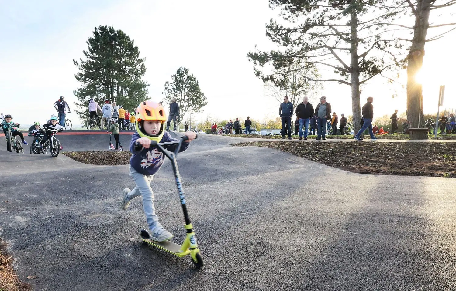 Ein Kind flitzt in Gaisbach mit seinem Tretroller über einen Pumptrack, der ähnlich wie eine Rollsportanlage in die Landschaft integriert wird. Drumherum gibt es Aufenthaltsflächen. ⇥