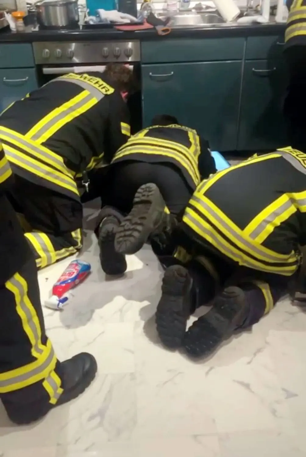 Knieend versuchen mehrere Feuerwehrmänner, die Katze hinter der Küchenzeile hervorzuholen. ⇥