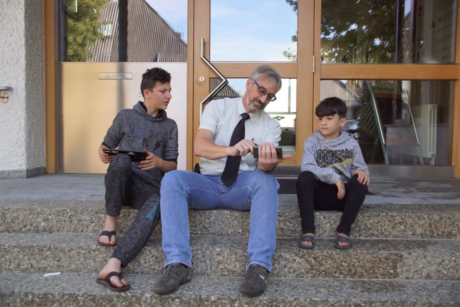 Karl-Friedrich Häcker sitzt vor dem Rathaus mit den Flüchtlingskindern Abbas und Adnan Al Fars, die das WLan im Rathaus nutzen.