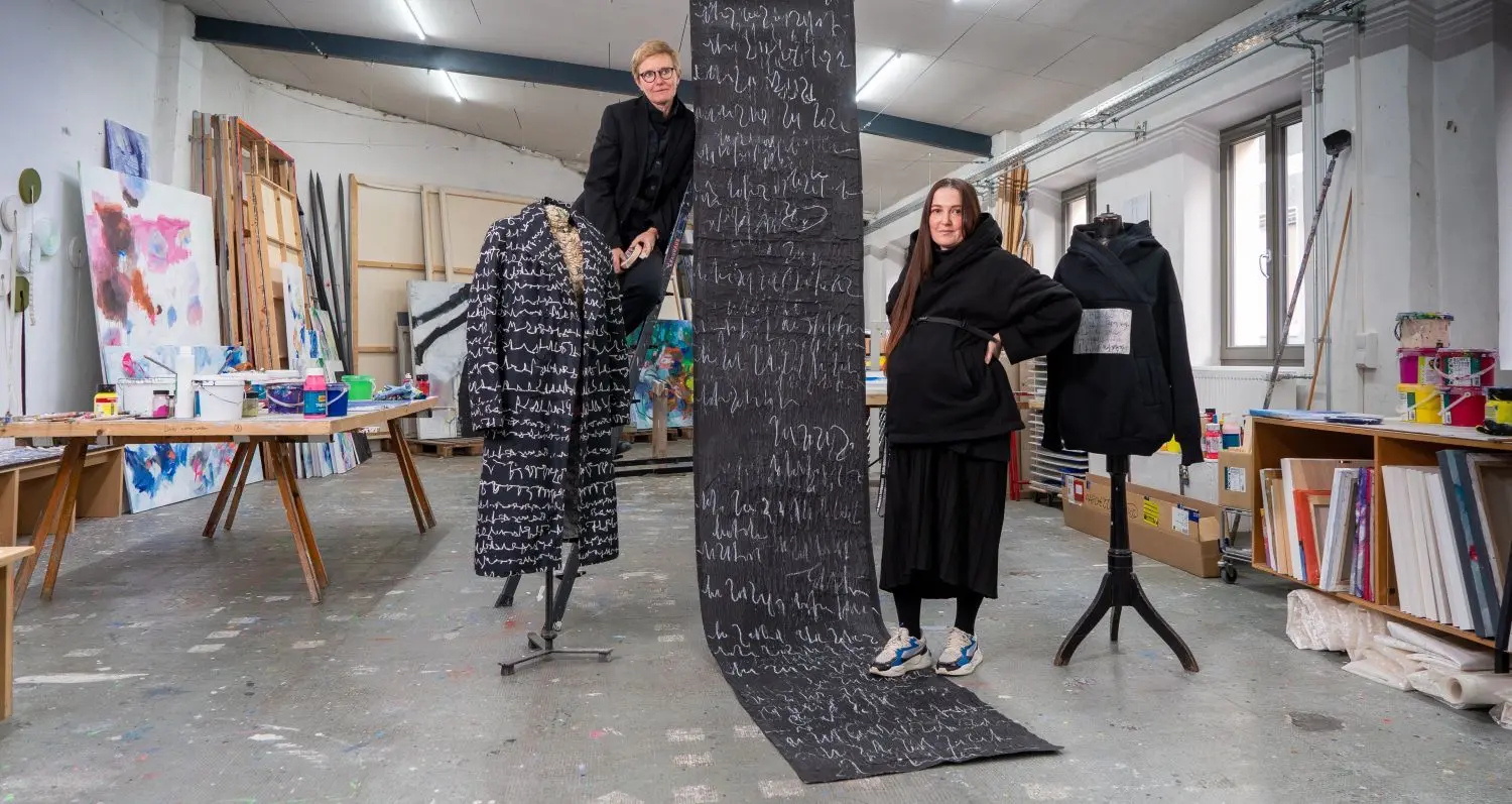 Kateryna Biakova mit Marianne Hollenstein (links) in deren Atelier. In der Mitte ein von der Ukrainerin designter Mantel, der auf einem Schrift-Kunstwerk der Ulmerin basiert.