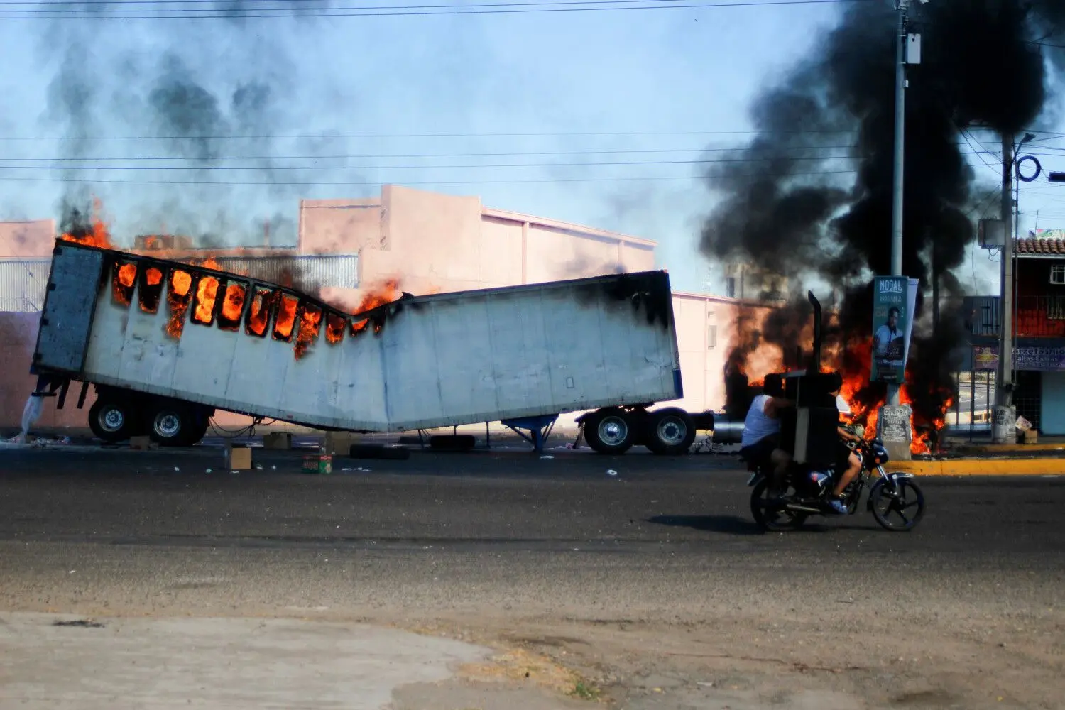 05.01.2023, Mexiko, Culiacan: Männer fahren auf einem Motorrad an einem brennenden Lastwagen in den Straßen von Culiacan im Bundesstaat Sinaloa vorbei. Nach der Festnahme eines Sohns des inhaftierten Drogenbosses Joaquín «El Chapo» Guzmán, Ovidio Guzmán, haben sich im Nordwesten von Mexiko mutmaßlichen Bandenmitgliedern und Sicherheitskräften über Stunden hinweg Kämpfe geliefert.