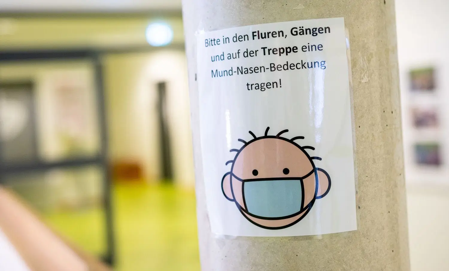 Ein Schild mit der Aufschrift "Bitte in den Fluren, Gängen und auf der Treppe eine Mund-Nasen-Bedeckung tragen!" ist an einer Säule eine Schule zu lesen.