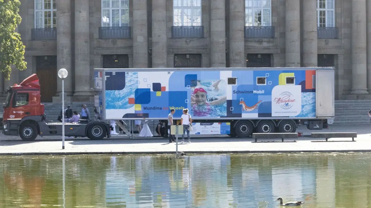 Etwa 13 Meter ist das Schwimm-Mobil lang. Zu Beginn macht es Station in Würzburg.
07.09.2022, Baden-Württemberg, Stuttgart: Ein ·Schwimm-Mobil· steht bei seiner Präsentation vor dem Operhaus, im Vordergrund ist der Eckensee zu sehen. Das Schwimm-Mobil «Wundine on Wheels» ist ein komplett ausgestattetes, mobiles Lehrschwimmbecken, das mit einem Sattelzug an den jeweiligen Einsatzort transportiert wird. Kinder im Alter von fünf bis acht Jahren können hier in Kleingruppen ihre ersten Erfahrungen beim Schwimmen machen. Foto: Marijan Murat/dpa +++ dpa-Bildfunk +++