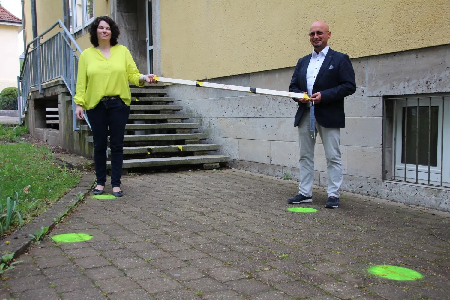 Schulleiter Michael Ullrich und die stellvertretende Schulleiterin Iris Heck mit der „Corona-Messlatte“ und Abstandsmarkierungen am Haupteingang der Leonhard-Sachs-Schule.⇥