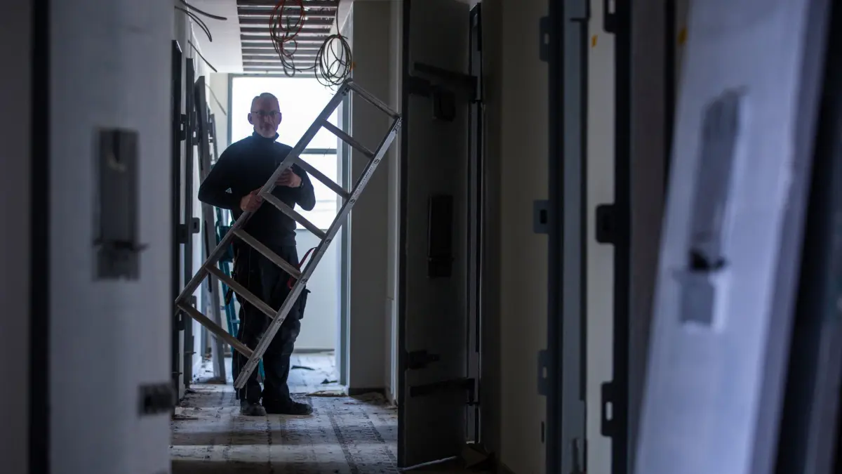 Aus Jugendgefängnis wird Hostel „Kittchen“: PRODUKTION - 20.03.2024, Mecklenburg-Vorpommern, Wismar: Handwerker arbeiten beim Umbau zum Hostel „Das Kittchen“ in einem Zellengang in der ehemalige Jugend-Arrestanstalt zwischen den originalen Haftraumtüren. Ein Übernachtungserlebnis der besonderen Art will ab Sommer ein Hostel in der Innenstadt bieten. Für rund 3,5 Millionen Euro wird die 2008 stillgelegte Einrichtung derzeit umgebaut. Etwa 100 Betten in 31 Zimmern sind geplant. Ein Teil der Hotelzimmer seien frühere Gefängniszellen - die kleinste messe ganze zwei mal dreieinhalb Meter. Mit einem Doppelstockbett können auf den acht Quadratmetern trotzdem zwei Gäste übernachten. (zu dpa: «Im «Kittchen» sind bald Zimmer frei - Herberge in Ex-Arrestanstalt») Foto: Jens Büttner/dpa +++ dpa-Bildfunk +++