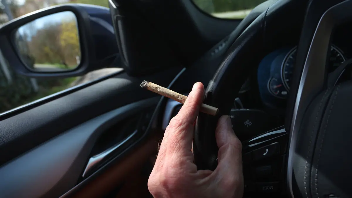 Vor der Cannabis–Legalisierung: ILLUSTRATION - 20.03.2024, Bayern, Kaufbeuren: Ein Mann sitzt mit einem Joint zwischen den Fingern am Steuer eines Autos. Der Bundesrat hatte am 22. März den Weg zur Teil-Legalisierung von Cannabis zum 1. April freigemacht. (zu dpa: «Innenministerin gegen höheren Cannabis-Grenzwert im Straßenverkehr») Foto: Karl-Josef Hildenbrand/dpa +++ dpa-Bildfunk +++