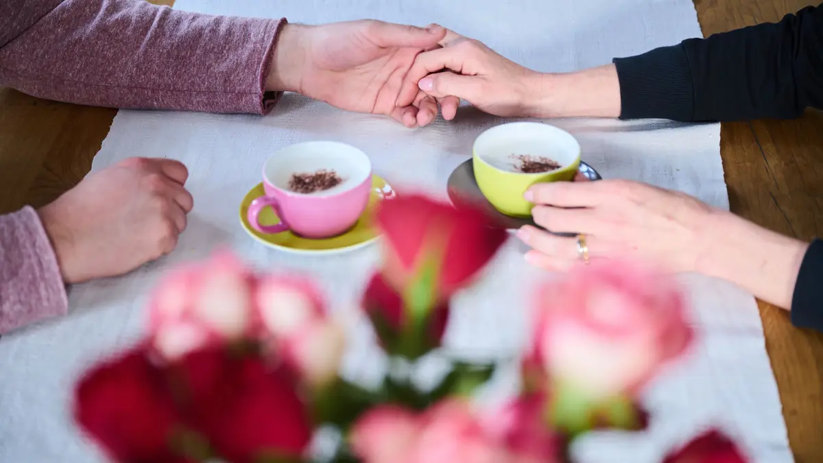 Ein Mann und eine Frau halten Händchen während Kaffeetassen und rote Rosen auf dem Tisch stehen. Am 14.02.2023 wird mit dem Valentinstag der Tag der Liebenden begangen. +++ dpa-Bildfunk +++