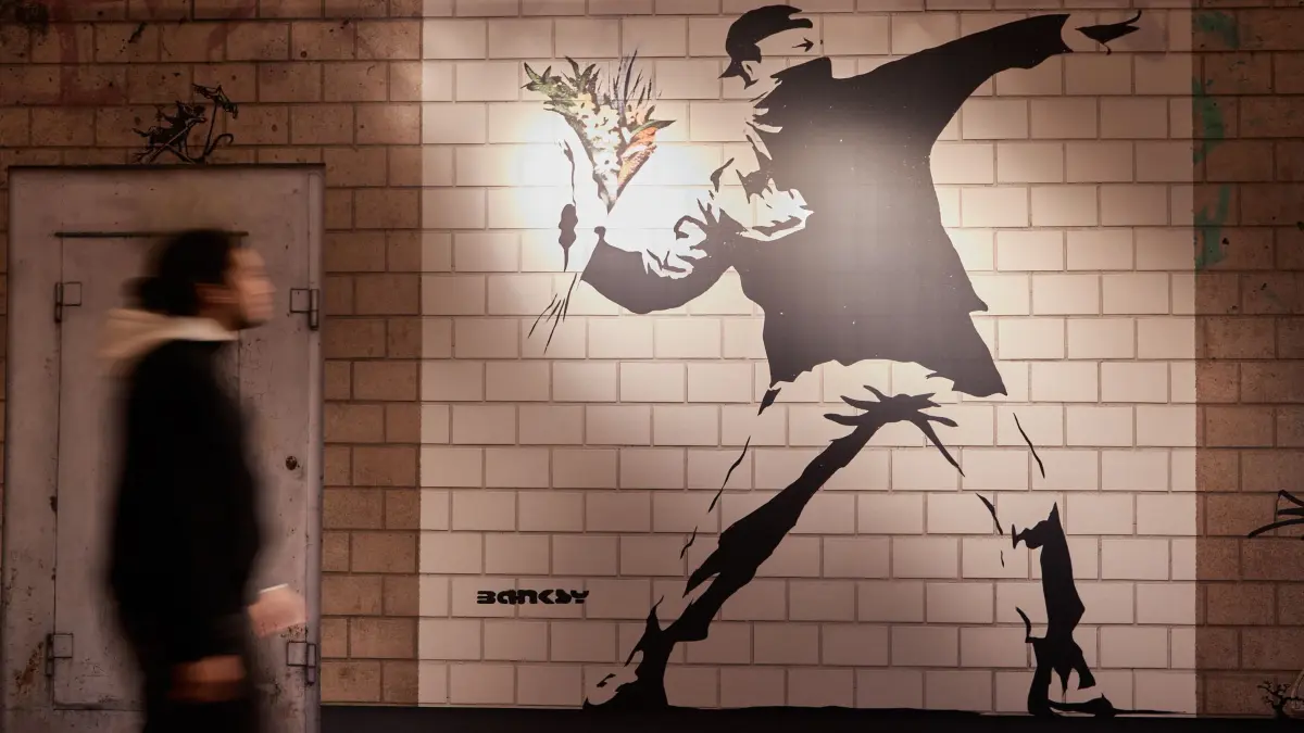 Ausstellung „The Mystery of Banksy - A Genius Mind“: ARCHIV - 02.06.2022, Hamburg: Ein Mitarbeiter betrachtet in der Ausstellung "The Mystery of Banksy - A Genius Mind" in der ehemaligen Galeria Kaufhof das Wandbild mit dem Titel "Flower Thrower". (zu dpa: «Banksy kommt zurück nach Hamburg») Foto: Georg Wendt/dpa +++ dpa-Bildfunk +++