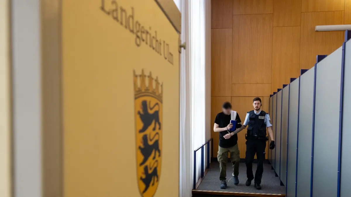Beginn Prozess wegen Geiselnahme in Ulm: 09.09.2024, Baden-Württemberg, Ulm: Ein Justizwachtmeister führt einen an Händen und Füßen gefesselten Mann in den Gerichtssaal. Dem Angeklagten wird vorgeworfen, Ende Januar 2024 insgesamt ein Dutzend Gäste und Mitarbeitende in einem Café in Ulm als Geiseln genommen zu haben. Der Soldat aus Nordrhein-Westfalen habe die Geiseln mit einer täuschend echten Pistolen-Attrappe bedroht und aufgefordert, die Polizei zu rufen. Sein Ziel sei gewesen, die einschreitenden Polizeibeamten während der laufenden Geiselnahme zu einem finalen «Rettungsschuss» zu zwingen. Wie von ihm erwartet, eröffneten die Polizisten das Feuer, woraufhin der Angeschuldigte getroffen und im Gesicht schwer verletzt wurde. Sämtliche Geiseln blieben unverletzt. Foto: Stefan Puchner/dpa - ACHTUNG: Person wurde aus rechtlichen Gründen gepixelt +++ dpa-Bildfunk +++