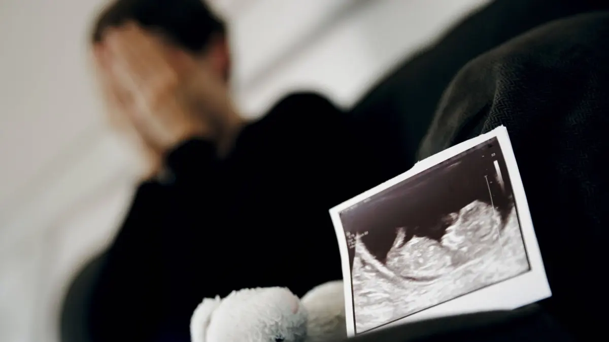 Symbolfoto: Gestellte Aufnahme zum Thema Fehlgeburt. Das Ultraschallbild eines ungeborenen Kindes steht neben einem Kuscheltier waehrend eine Frau im Hintergrund ihr Gesicht in ihren Haenden verbirgt. Berlin, 22.08.2023. Berlin Deutschland *** Symbol photo Posed image on the subject of miscarriage The ultrasound image of an unborn child stands next to a cuddly toy while a woman in the background hides her face in her hands Berlin, 22 08 2023 Berlin Germany Copyright: xThomasxTrutschelx
