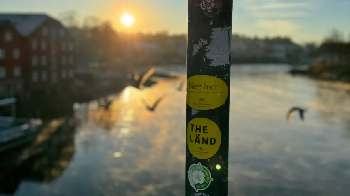An einem Pfahl auf einer Brücke kleben Aufkleber mit der Aufschrift „Nett hier“ und „The Länd“. Die Sticker sind Teil der Kampagne „The Länd“ des Landes Baden-Württemberg. Das Land möchte am liebsten, dass Menschen die Sticker nur noch fotografieren, statt zu kleben.