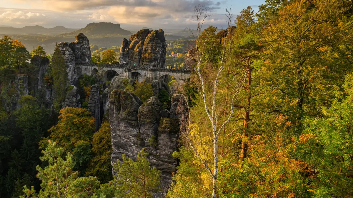 Sonnenlicht fällt am Morgen auf die herbstlich gefärbten Bäume im Nationalpark Sächsische Schweiz nahe der 1851 gebauten Basteibrücke. Im Hintergrund ist der Lilienstein zu sehen. +++ dpa-Bildfunk +++