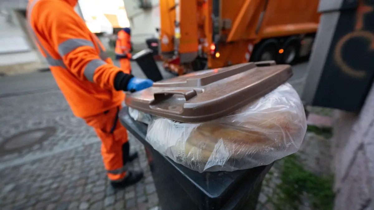 KI-unterstützte Biomüll-Abfuhr in Reutlingen: ARCHIV - 19.03.2025, Baden-Württemberg, Reutlingen: Eine mit Plastikmüll und Fremdstoffen falsch befüllte Biomülltonne steht in der Innenstadt, dahinter leeren Mitarbeiter der Technischen Betriebsdienste Reutlingen (TBR) Biomülltonnen. (zu dpa: «Wie Entsorger mit dem neuen Grenzwert für Biomüll umgehen») Foto: Marijan Murat/dpa +++ dpa-Bildfunk +++