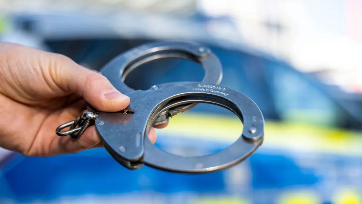 Haftbefehl - Symbolbild: ILLUSTRATION - 28.09.2021, Nordrhein-Westfalen, Gütersloh: Eine Hand hält Handschellen vor einen Streifenwagen der Polizei. (zu dpa: «Nach Messerattacke in München: Verdächtiger in U-Haft») Foto: David Inderlied/dpa +++ dpa-Bildfunk +++