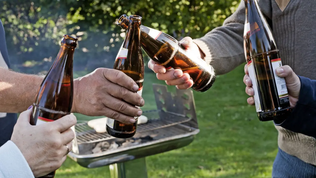 Vier Leute stoßen draußen vor einem Grill mit Bierflaschen an: ARCHIV - Ein Kühlschrank im Garten sorgt für kühle Getränke. Am richtigen Platz ist dafür dann auch nicht massig Energie nötig. (zu dpa: «Sommerparty: Ein Schattenplätzchen für den Kühlschrank») Foto: Monique Wüstenhagen/dpa-tmn - Honorarfrei nur für Bezieher des dpa-Themendienstes +++ dpa-Themendienst +++