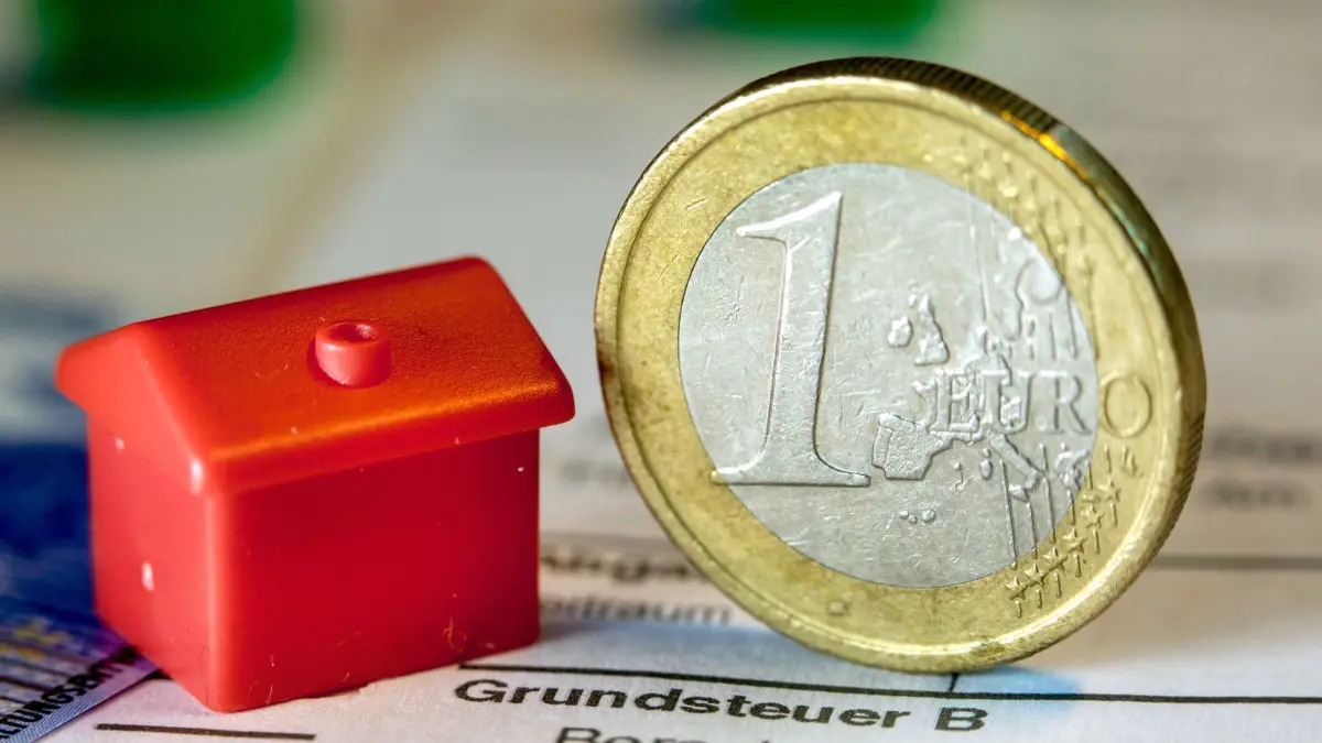ARCHIV - 30.01.2019, Mecklenburg-Vorpommern, Schwerin: Illustration - Eine Euro-Geldmünze sowie ein Spielzeughaus stehen auf einem Abgabenbescheid für die Entrichtung der Grundsteuer. (zu dpa: «Grund- und Gewerbesteuer bleibt in Sachsen-Anhalt stabil»). Foto: Jens Büttner/dpa +++ dpa-Bildfunk +++