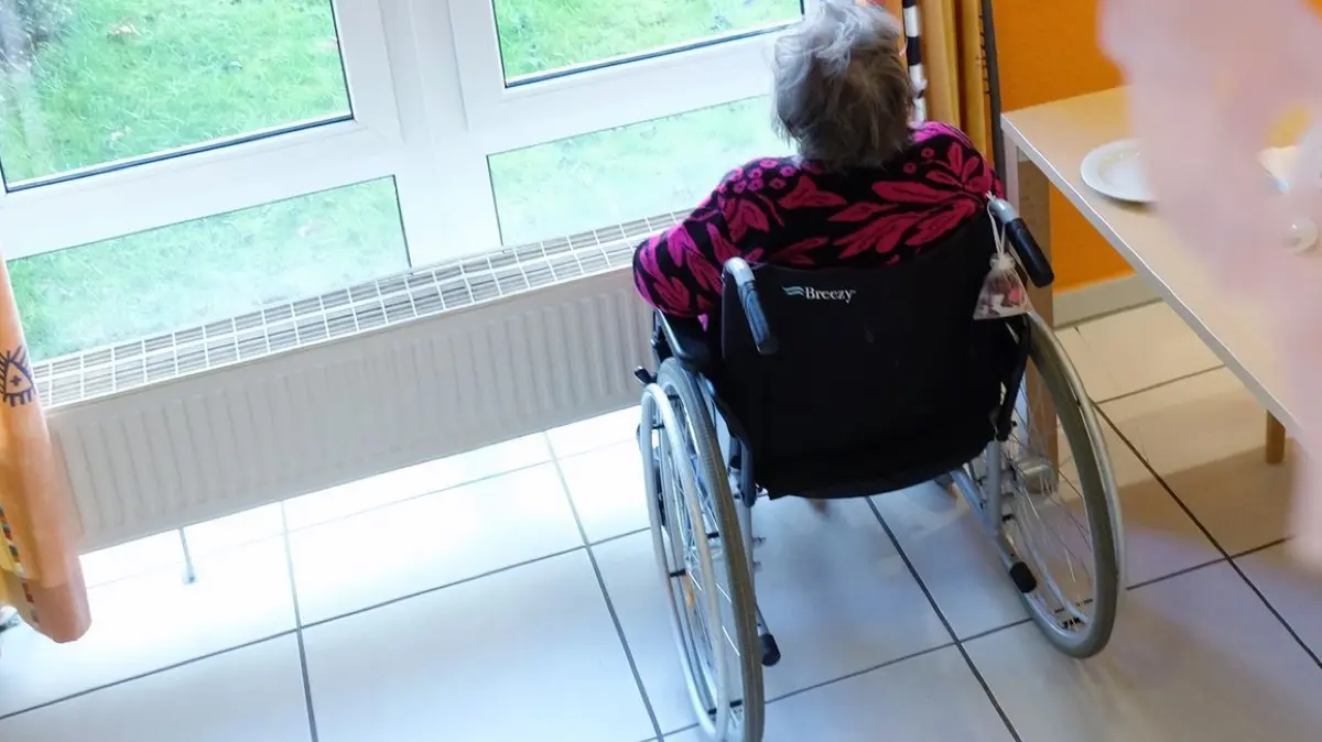 Einsamkeit im Alter: ARCHIV - 20.01.2022, Sachsen, Delitzsch: Die Bewohnerin eines Pflegeheimes mit Faschingsdekoration im Vordergrund sitz in einem Rollstuhl und blickt aus dem Fenster. Die Gesundheitsministerin des Landes Sachsen, Köpping (SPD), will sich über das Projekt «Kiko - Chatbot für Seniorinnen und Senioren» informieren. (zu dpa: «Köpping besucht Projekt zu KI gegen Einsamkeit im Alter») Foto: Sebastian Willnow/dpa-Zentralbild/dpa +++ dpa-Bildfunk +++