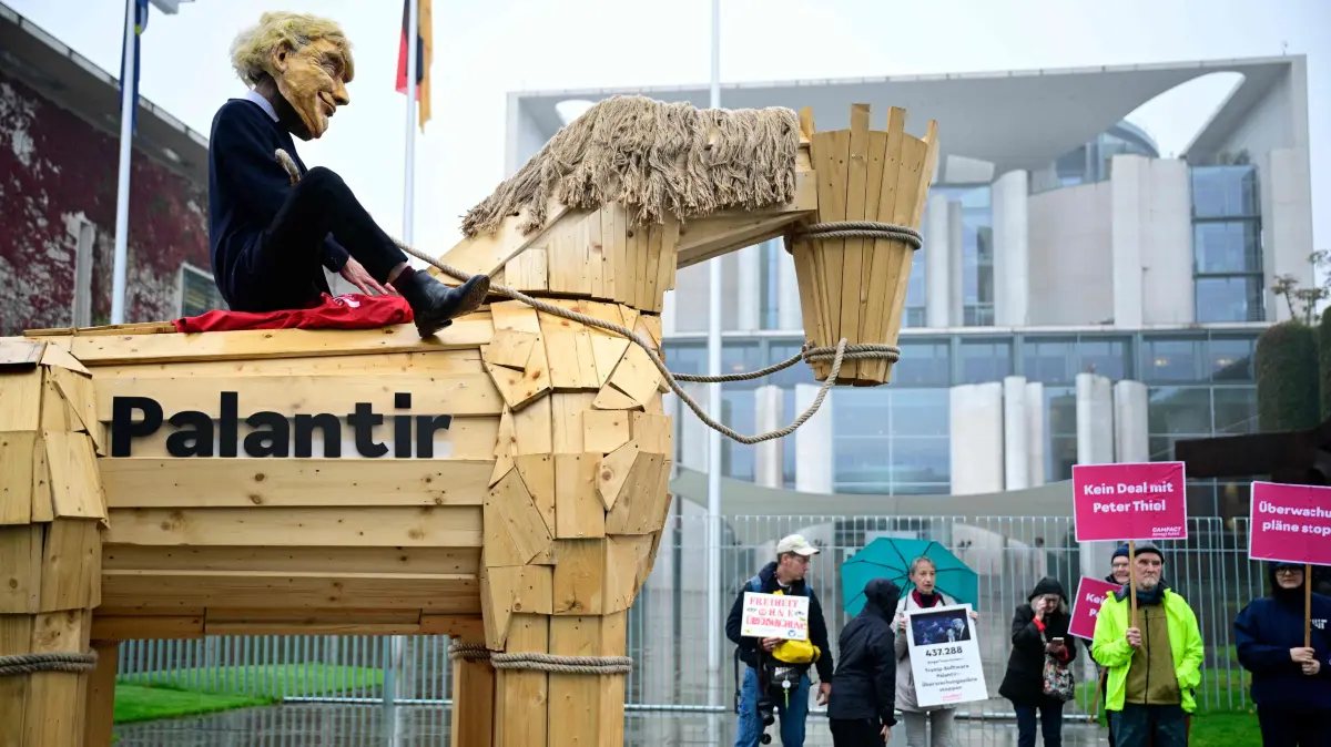 An activist of the group Campact! dressed as US President Donald Trump (L) sits on a wooden horse symbolising a Trojan Horse with the lettering "Palantir" in front of the Chancellery in Berlin, where is taking place the German cabinet's weekly meeting on October 8, 2025. Activists protested against the German Interior Ministry's intention to pave the way for the nationwide use of the data mining Palantir software, that was developed by a company co-founded and chaired by Trump supporter Peter Thiel. (Photo by Tobias SCHWARZ / AFP)