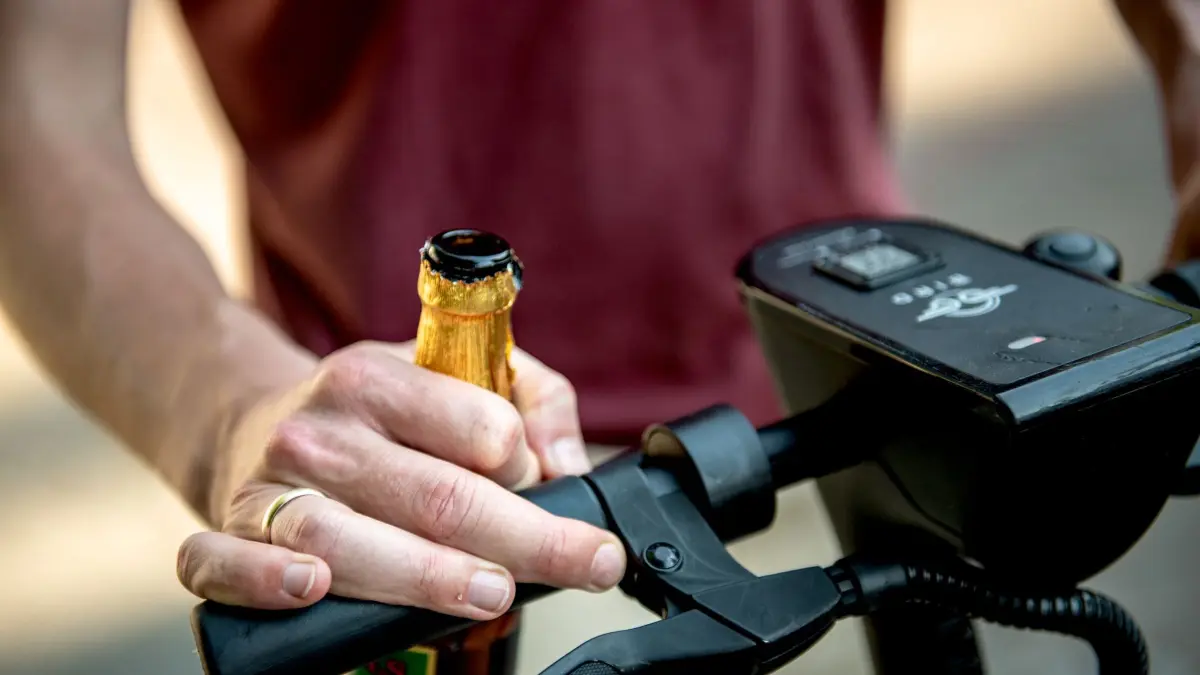 Ein Mann hält ein Bier und einen E-Scooter fest: ILLUSTRATION - Alkohol am Roller-Steuer: Für E-Scooter gilt dieselbe Promillegrenze wie fürs Auto. (zu dpa: «Balanceakt E-Scooter: Sicherheit statt Risiko») Foto: Zacharie Scheurer/dpa-tmn - Honorarfrei nur für Bezieher des dpa-Themendienstes +++ dpa-Themendienst +++