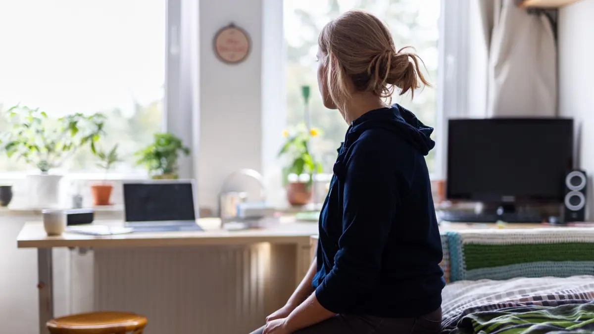 Eine Studentin sitzt auf dem Bett in ihrem WG-Zimmer und schaut aus dem Fenster.(gestellte Szene)
