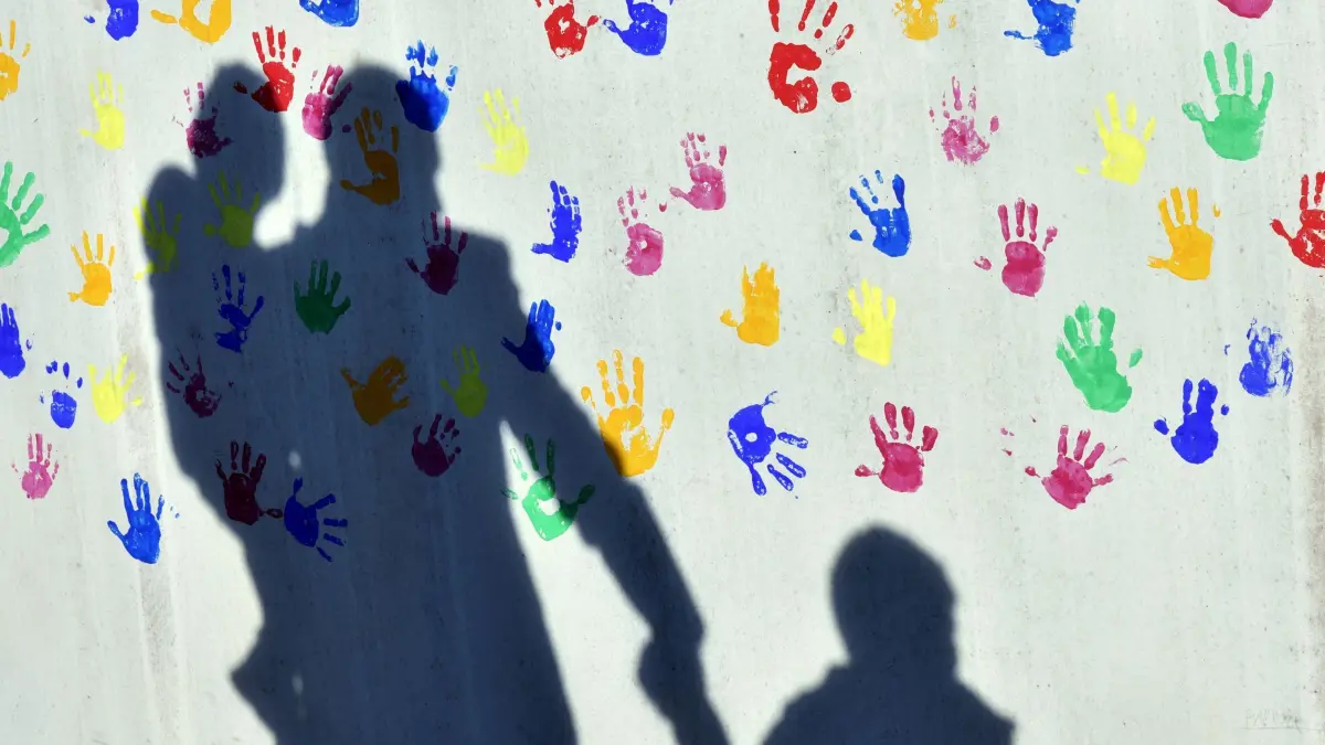Pflegefamilie: ARCHIV - 08.04.2015, Bayern, München: Ein Mann mit einem Kind auf dem Arm und einem an der Hand wirft einen Schatten auf eine mit bunten Handabdrücken bemalte Wand einer Kindertagesstätte. (zu dpa: «Pflegefamilien gesucht – Kinder warten in Hessen auf Zuhause») Foto: Peter Kneffel/dpa +++ dpa-Bildfunk +++