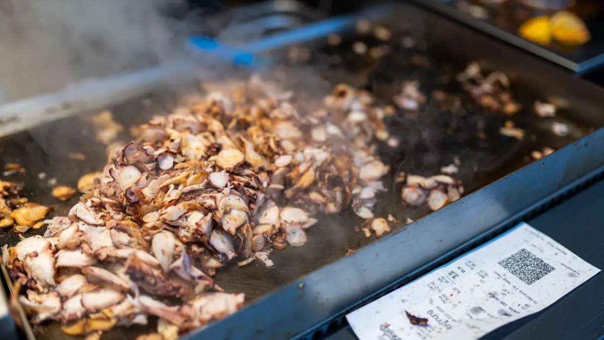 Calamari-Kebab: PRODUKTION - 07.11.2025, Baden-Württemberg, Karlsruhe: Abgeschnittenes und gebratenes Fleisch liegt auf einer Grillplatte neben einem Calamari-Kebabspiess im Restaurant «Pescobar». Nach der ersten Filiale in London hat ein rumänischer Meeresfrüchte-Anbieter nun seine zweite Filiale mit Calamari-Kebab in Karlsruhe eröffnet. (zu dpa: «Neuer Döner-Hype? Tentakel am Spieß sorgt für Wirbel») Foto: Philipp von Ditfurth/dpa +++ dpa-Bildfunk +++