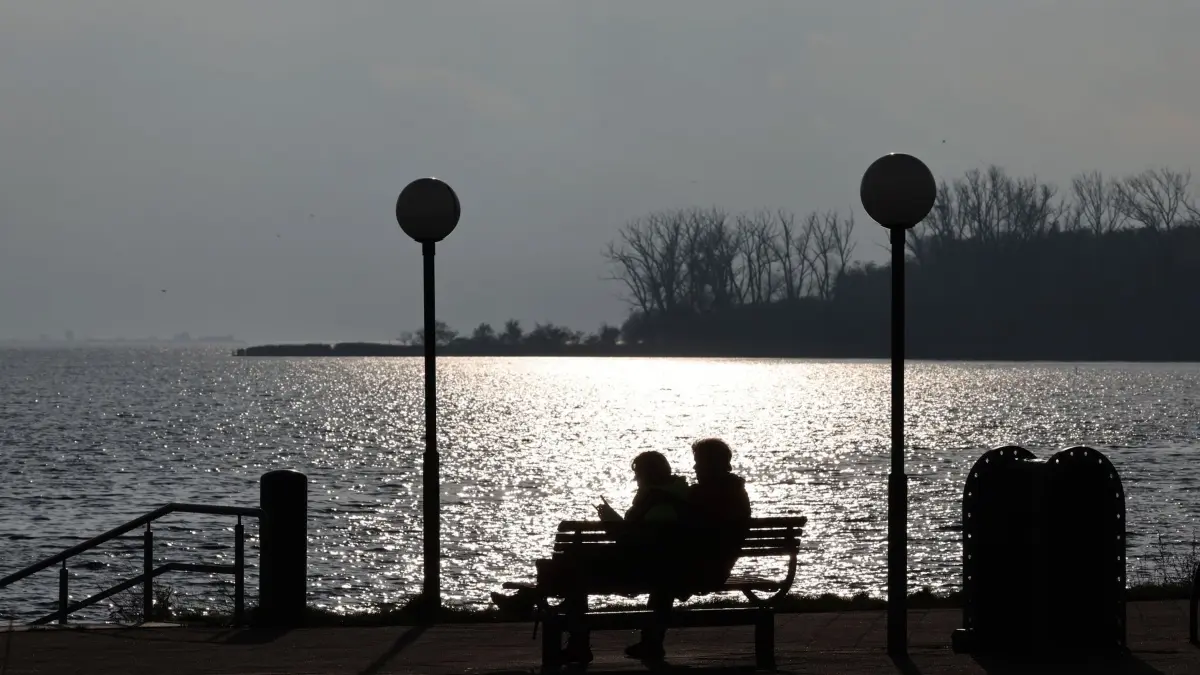 Ruhiges Herbstwetter im Norden: 11.11.2025, Mecklenburg-Vorpommern, Rerik: Spaziergänger sitzen im Hafen am Salzhaff auf einer Bank, während dunkle Wolken aufgezogen sind. Die Sonne zeigt sich nur kurz und leuchtet auf dem Wasser. Foto: Bernd Wüstneck/dpa +++ dpa-Bildfunk +++