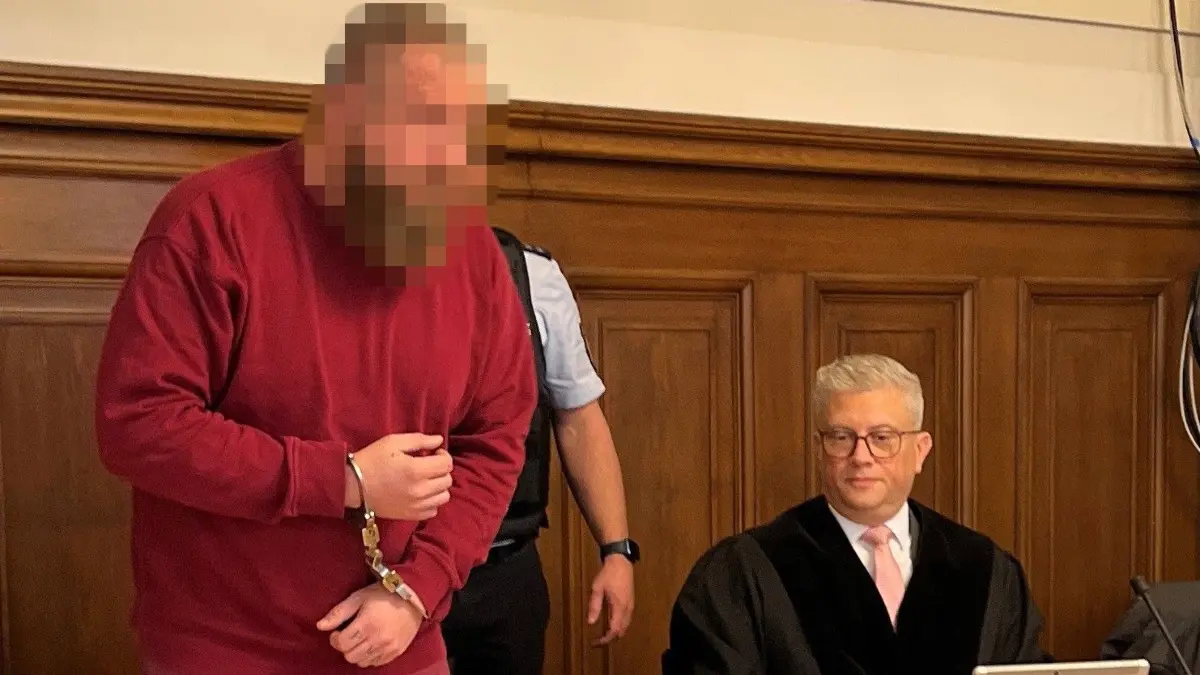 In Handschellen und mit Fußfesseln wird er Angeklagte von zwei Wachtmeistern auf den Platz neben seinem Anwalt geführt.
