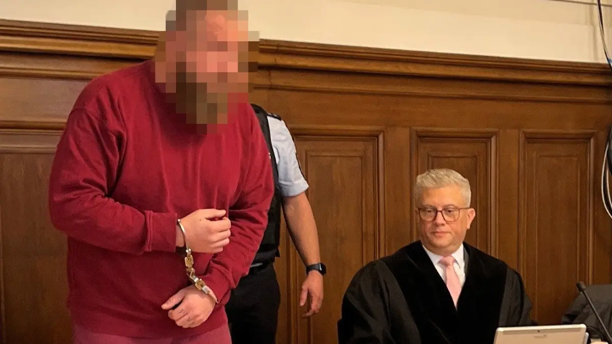In Handschellen und mit Fußfesseln wird er Angeklagte von zwei Wachtmeistern auf den Platz neben seinem Anwalt geführt.