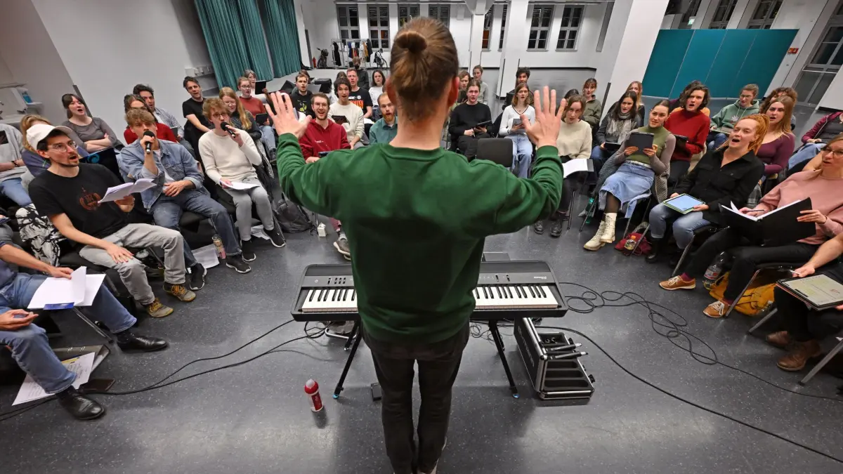 Zulauf bei Thüringer Chören: PRODUKTION - 19.11.2025, Thüringen, Jena: Maximilian Lörzer, Chorleiter des Psycho-Chors der FSU Jena, dirigiert die Chormitglieder bei einer Probe in der Aula der Ernst-Abbe-Hochschule. Das gemeinsame Singen in Chören ist in Thüringen beliebt. Der Thüringer Chorverband hatte Ende 2024 nach eigenen Angaben 259 Mitgliedschöre mit rund 6.500 Aktiven. Die Gesamtzahl aller Chöre in Thüringen ist nicht bekannt, weil viele Schul- und Kirchenchöre nicht zentral erfasst werden. (zu dpa: ««Die Leute brennen für das Singen» - Chöre beliebt») Foto: Martin Schutt/dpa +++ dpa-Bildfunk +++