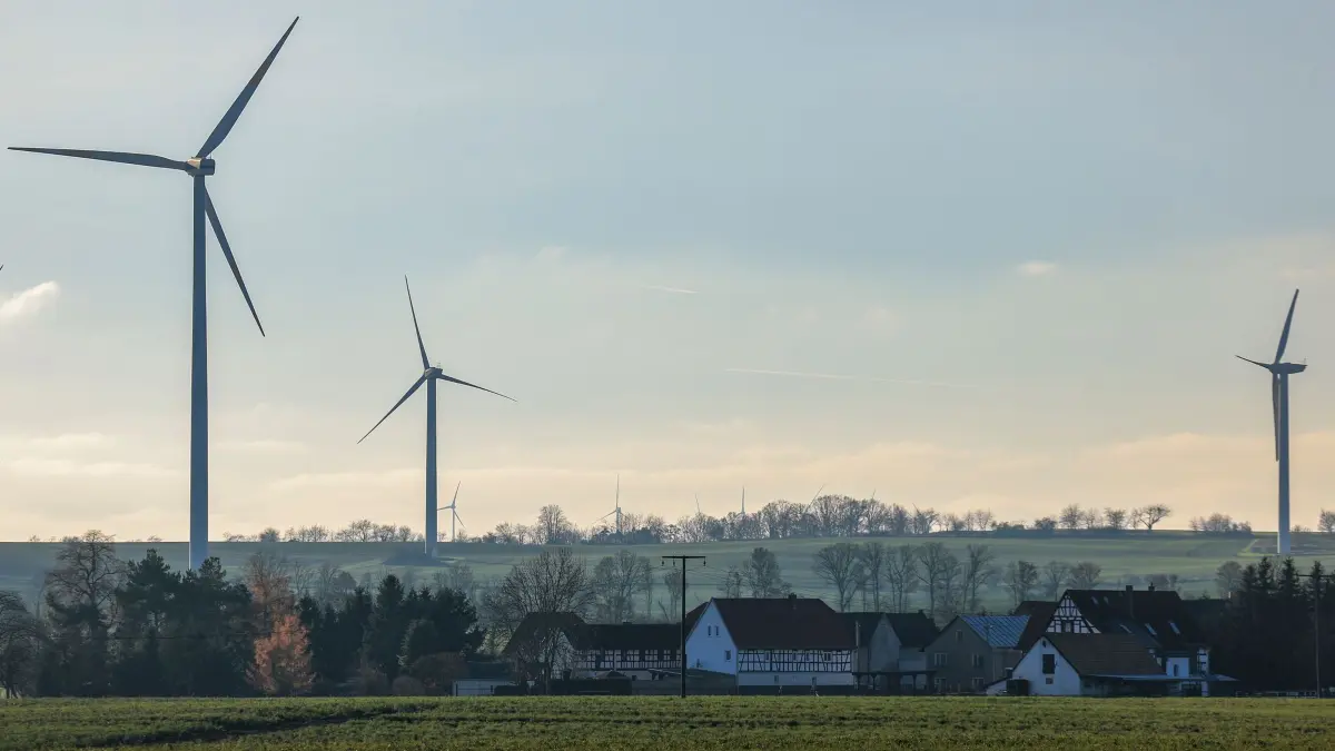 Windräder hinter Dorf: 04.12.2025, Thüringen, Neubraunshain: Mehrere Windkraftanlagen stehen hinter dem kleinen Ort. In Thüringen gelten für Windräder seit 2024 Mindestabstände zu Wohngebieten von 1000 Metern. Foto: Jan Woitas/dpa - Honorarfrei nur für Bezieher des Dienstes ZB-Funkregio Ost +++ ZB-FUNKREGIO OST +++