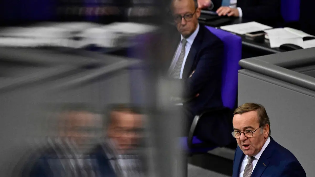 German Chancellor Friedrich Merz (background) listens as German Defence Minister Boris Pistorius addresses delegates during a plenary session at the Bundestag, Germany's lower house of parliament in Berlin on December 5, 2025. German MP's are voting on a controversial pension package and a new military service plan. (Photo by John MACDOUGALL / AFP)