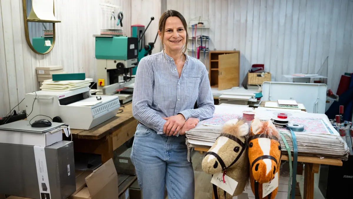 100 Jahre Buchbinderei Helmrich: Antje Helmrich in dem hinteren Teil des Ladens, wo die Bücher entstehen. Links hinter ihr stehen eine Bohrmaschine (grün), um Löcher in Pappe zu machen sowie links daneben eine Spiralmaschine, die die Spiralen eines Albums schließt.