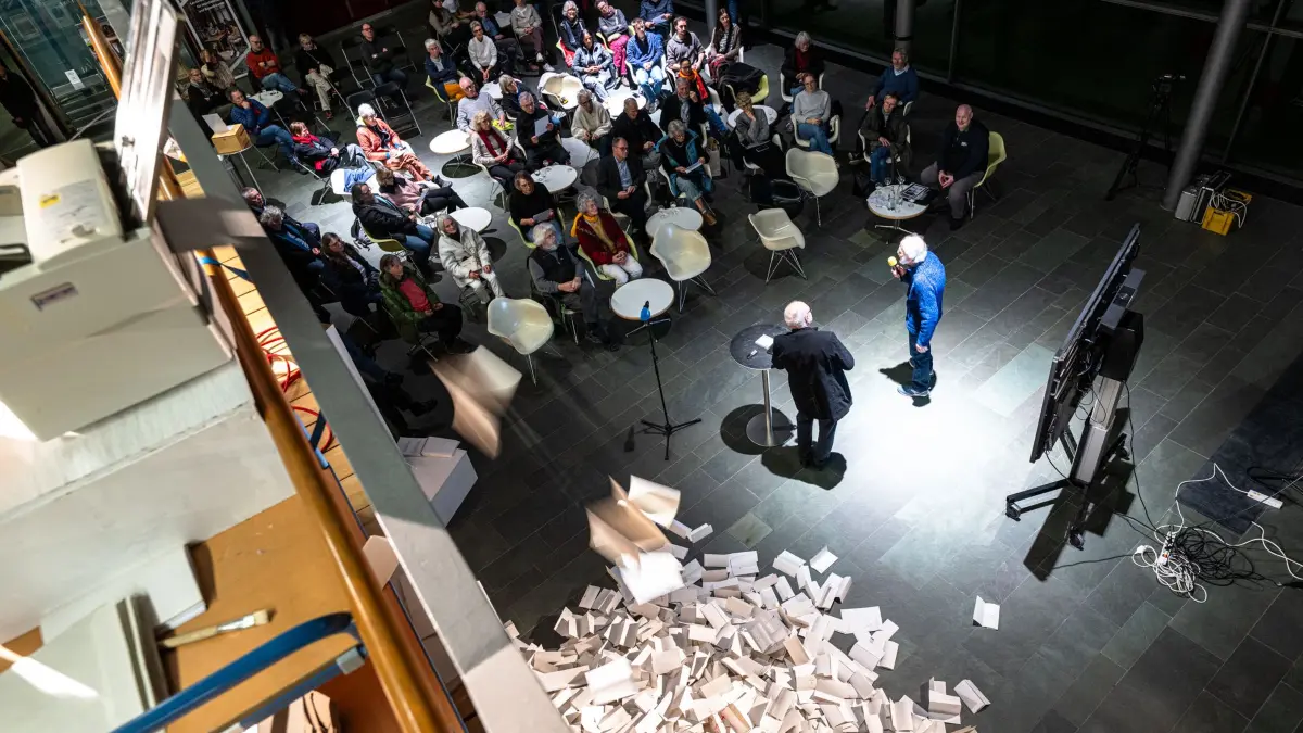 Kunstaktion im Landratsamt Tübingen zu 85 Jahre Grafeneck: 10.654 Gedenkbriefe spuckt die Falzmaschine (links) von der Empore in das Foyer des Landratamts, während der Künstler Jochen Meyder (im blauen Pullover) mit Kreisarchivar Wolfgang Sannwald über seine Kunstaktion und Grafeneck spricht.