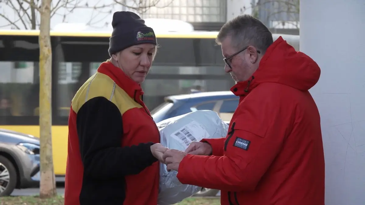 Unterwegs mit einem Paketboten im Weihnachtsstress: PRODUKTION - 13.12.2025, Berlin: Kerstin Klein (l), Paketbotin der Deutschen Post DHL, die eine Mütze mit dem Griesgram Grinch und dem Aufdruck «MIMIMI - Halts Maul!» trägt, übergibt ein Paket an Bernd Wendland. Der Hausbewohner versucht die Paketbotin zu entlasten, indem er auch für Nachbarn Pakete annimmt. In der Vorweihnachtszeit rechnen Zusteller mit deutlich mehr Sendungen - und einem entsprechend höheren Arbeitsdruck. Foto: Manuel Genolet/dpa - ACHTUNG: Nur zur redaktionellen Verwendung im Zusammenhang mit der aktuellen Berichterstattung und nur mit vollständiger Nennung des vorstehenden Credits +++ dpa-Bildfunk +++