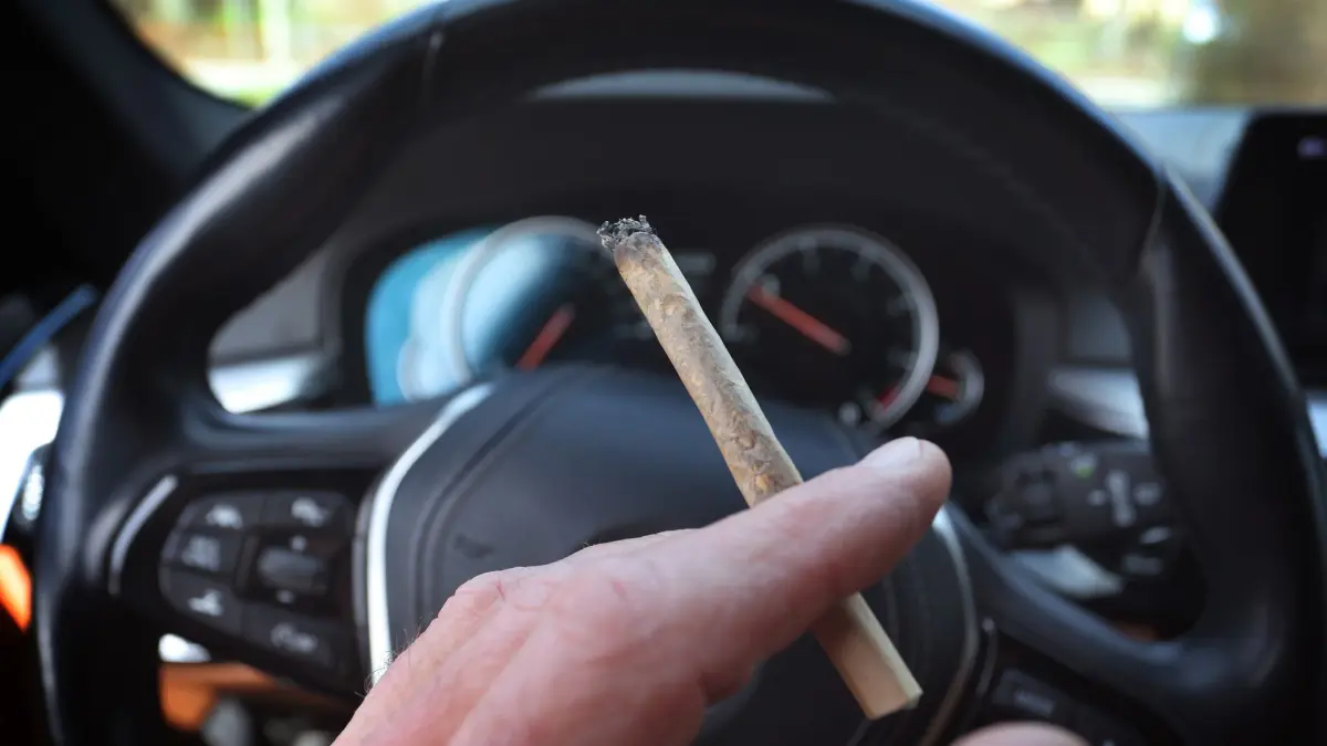 Cannabis am Steuer: ILLUSTRATION - 20.03.2024, Bayern, Kaufbeuren: Ein Mann sitzt mit einem Joint zwischen den Fingern am Steuer eines Autos. (zu dpa: «Innenminister zieht Bilanz zu Kontrollaktion der Polizei») Foto: Karl-Josef Hildenbrand/dpa +++ dpa-Bildfunk +++