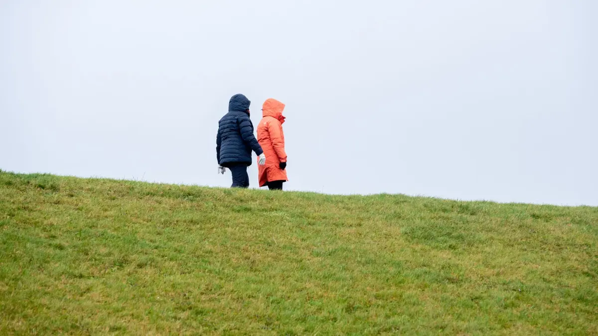 Wetter in Niedersachsen - Cuxhaven: 01.01.2026, Niedersachsen, Cuxhaven: Zwei Frauen gehen bei regnerischem und stürmischem Wetter auf einem Deich im Ortsteil Döse entlang. Foto: Hauke-Christian Dittrich/dpa +++ dpa-Bildfunk +++