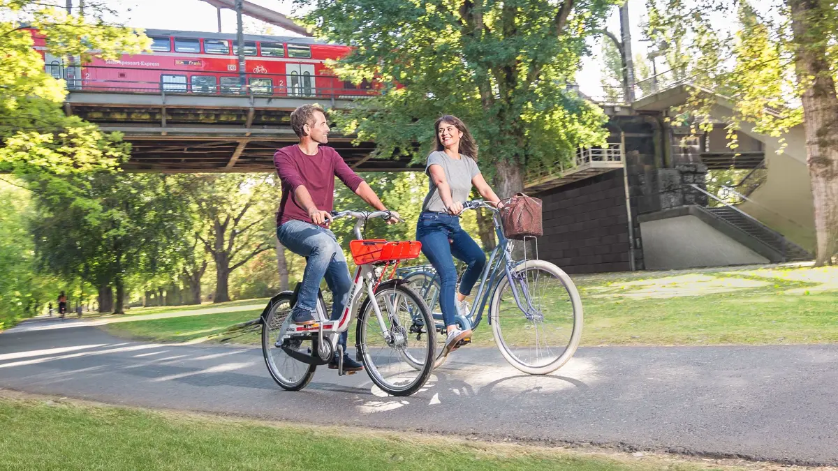 Anschlussmobilität mit dem Fahrrad: Pärchen fährt auf privatem Rad und Call a Bike durch einen Park. Im Hintergrund eine Regionalbahn von DB Regio