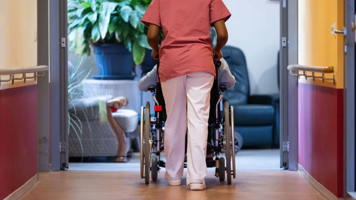 Eine Pflegekraft schiebt einen Pflegebedürftigen im Rollstuhl: ILLUSTRATION - Wohngeld, Pflegewohngeld, Hilfe zur Pflege: Wird das Geld knapp, können Pflegebedürftige im Heim Unterstützung bekommen. (zu dpa: «Geld reicht nicht mehr fürs Pflegeheim - was tun?») Foto: Marijan Murat/dpa/dpa-mag - Honorarfrei nur für Bezieher des Dienstes dpa-Magazin +++ dpa-Magazin +++