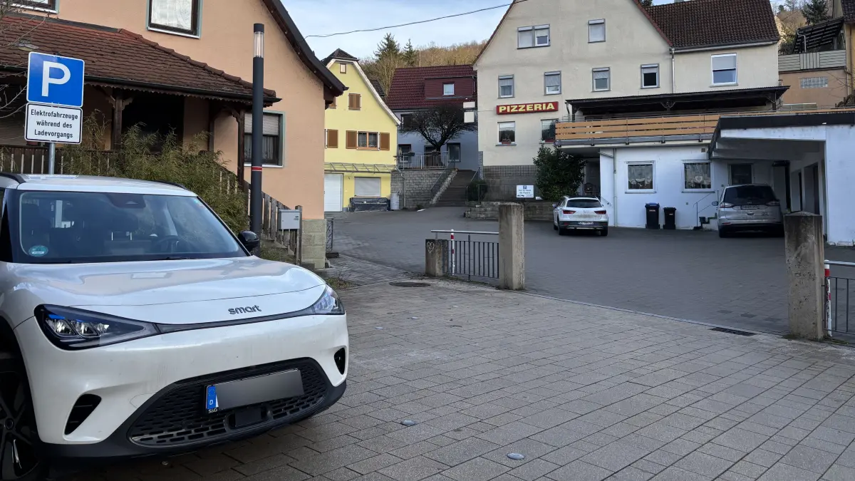 Zwei klar gekennzeichnete Parkplätze für E-Fahrzeuge mit Ladestation gibt es auf dem Braunsbacher Marktplatz. Sie grenzen an eine Einfahrt zur Pizzeria O Sole Mio und weiteren Gebäuden.