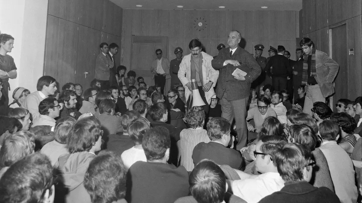 Studenten besetzen das Tübinger Rathaus, 25. Mai 1968.
Nachdem es Oberbürgermeister Hans Gmelin abgelehnt hatte, mit einer in der Haaggasse versammelten „Corona“ zu diskutieren, drangen gegen 10.15 Uhr etwa 300 Studenten, angeführt von Mitgliedern des Sozialistischen Deutschen Studentenbundes (SDS), gegen den Widerstand von drei Polizeibeamten in das Rathaus ein. Womit, wie das Schwäbische Tagblatt zu berichten wusste, das Tübinger Rathaus in seiner über 500jährigen Geschichte „sein erstes Go-in“ erlebte. Was ihnen Tags zuvor vom Amt für öffentliche Ordnung verweigert worden war, wollten sie nun vom Oberbürgermeister genehmigt bekommen: einen Lautsprecherwagen, mit dem sie darüber aufklären wollten, wie sich die geplanten Notstandsgesetze auf den einzelnen Bürger auswirken würden. Gmelin (rechts im Bild stehend) zeigte sich im Laufe der erzwungenen, nahezu dreistündigen Diskussion kompromissbereit. Er wollte die Genehmigung jedoch örtlich und zeitlich beschränken, um den Verkehr während der Hauptzeiten nicht zu behindern.
Daraufhin beschloss eine knappe Mehrheit der Studenten, den Lautsprecherwagen auch ohne Genehmigung an den drei Tagen vor der Abstimmung im Bundestag ohne zeitliche und örtliche Beschränkung einzusetzen. Der Wagen wurde durch die Stadt nicht behindert; Oberbürgermeister und Mitglieder des Gemeinderats waren sich einig, dass ein massiver Polizeieinsatz nicht zu vertreten sei.
Versöhnlicher Schluss: Nach dem „Go-in“ war „das schöne neue Treppenhaus des Rathauses“... übersät von Kippen und Pfeifenasche. Empört wandte sich die offenbar herbeigeeilte Gattin des Oberbürgermeisters an die Studenten: „So nun gehen Sie, und den Dreck lassen Sie liegen!“ Aber siehe da: Fünf Studenten der schwäbischen Universitätsstadt „verlangten nach Besen und Kutterschaufel und halfen dem Hausmeister bei der Beseitigung ihrer Hinterlassenschaft“.
Studentenbewegung
Stadtarchiv Tübingen, D 170/12479