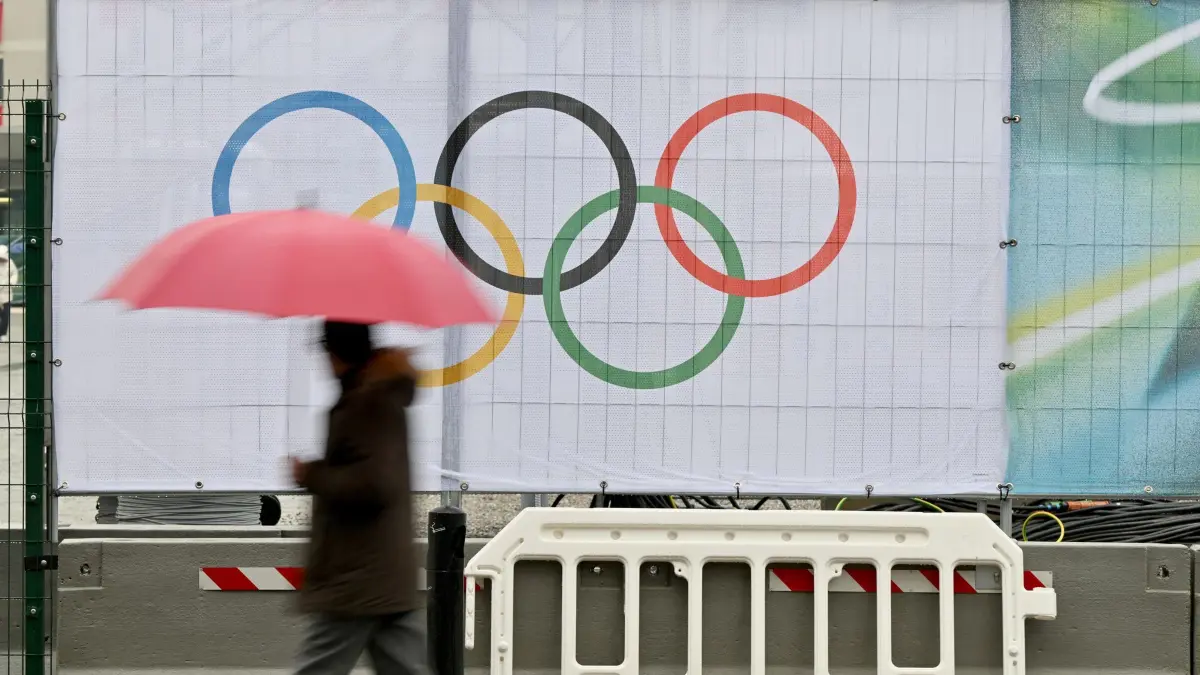 Olympische Winterspiele 2026: 04.02.2026, Italien, Mailand: Olympia, Olympische Winterspiele Mailand Cortina 2026, Ein Mann mit Regenschirm läuft an einem Sichtschutz mit den olympischen Ringen vorbei. Foto: Peter Kneffel/dpa +++ dpa-Bildfunk +++