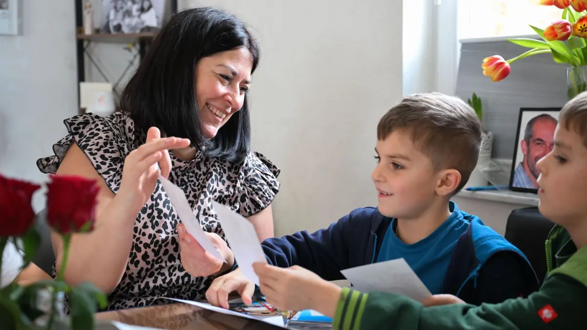 Grabsteine mit Briefschlitz: PRODUKTION - 17.02.2026, Baden-Württemberg, Bad Wildbad: Christina Schmid (l) schaut sich mit den Zwillingen Theo und Tom gemeinsame Familienfotos mit dem Familienvater an. Der Vater starb 2023 an Krebs. Auf seinem Grab steht ein Grabstein mit Briefschlitz, in den die Familie Briefe einwerfen kann. Foto: Katharina Kausche/dpa - ACHTUNG: Nur zur redaktionellen Verwendung im Zusammenhang mit der aktuellen Berichterstattung und nur mit vollständiger Nennung des vorstehenden Credits +++ dpa-Bildfunk +++