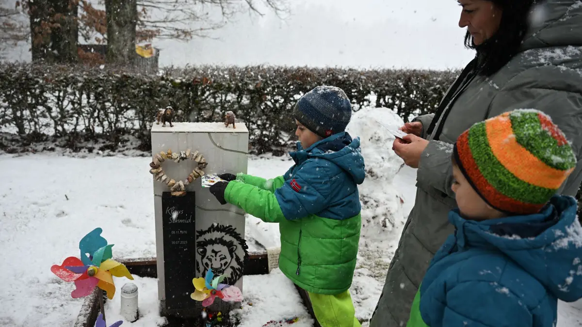 Grabsteine mit Briefschlitz: PRODUKTION - 17.02.2026, Baden-Württemberg, Bad Wildbad: Tom wirft einen Brief für seinen Vater in den Briefschlitz an seinem Grabstein. Neben ihm stehen seine Mutter Christina Schmid und sein Bruder Theo. Der Familienvater starb 2023 an Krebs. Auf seinem Grab steht ein Grabstein mit Briefschlitz, in den die Familie Briefe einwerfen kann. Foto: Katharina Kausche/dpa - ACHTUNG: Nur zur redaktionellen Verwendung im Zusammenhang mit der aktuellen Berichterstattung und nur mit vollständiger Nennung des vorstehenden Credits +++ dpa-Bildfunk +++