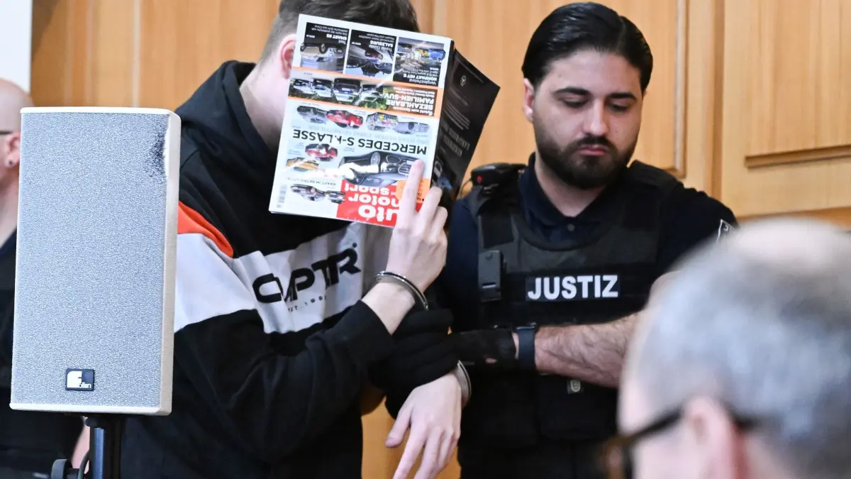Prozessbeginn nach gewaltsamen Tod auf Supermarkt-Parkplatz: 11.03.2026, Baden-Württemberg, Heilbronn: Ein Angeklagter (l) wird vor dem Beginn seines Prozesses mit einer Zeitung vor dem Gesicht in den Gerichtssaal geführt. Rechts steht sein Verteidiger Andreas Kugel. Der zur Tatzeit 18-jährige Angeklagte soll im September 2025 auf dem Parkplatz eines Supermarktes in Niedernhall mit seinem Auto einen 12-jährigen Jungen, der mit dem Fahrrad vorausfuhr, zielgerichtet angefahren haben. Die Anklage geht von Mord aus niedrigen Beweggründen aus. Foto: Bernd Weißbrod/dpa +++ dpa-Bildfunk +++
