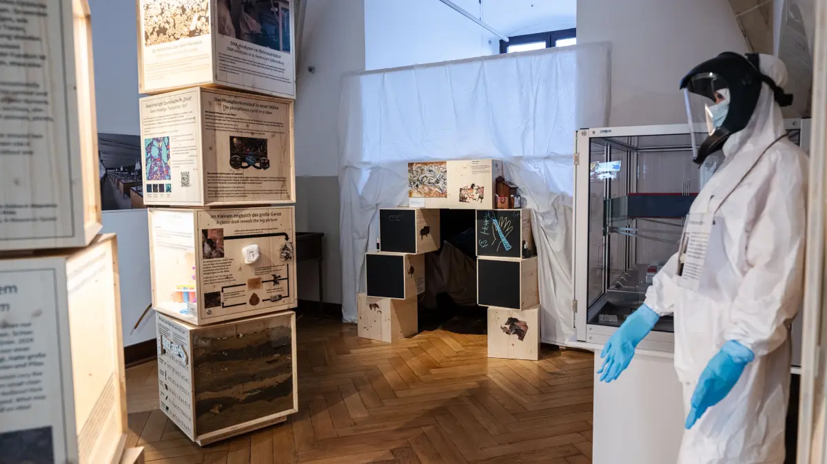 Ausstellung im MUT "Dirty Science – Höhlensedimente als Archive der Vergangenheit", Blick in die Ausstellung mit Höhle für Kinder (hinten), Reinraum-Puppe (rechts) und Holz-Modulen mit Texten, Bildern und Ausstellungsstücken (links)