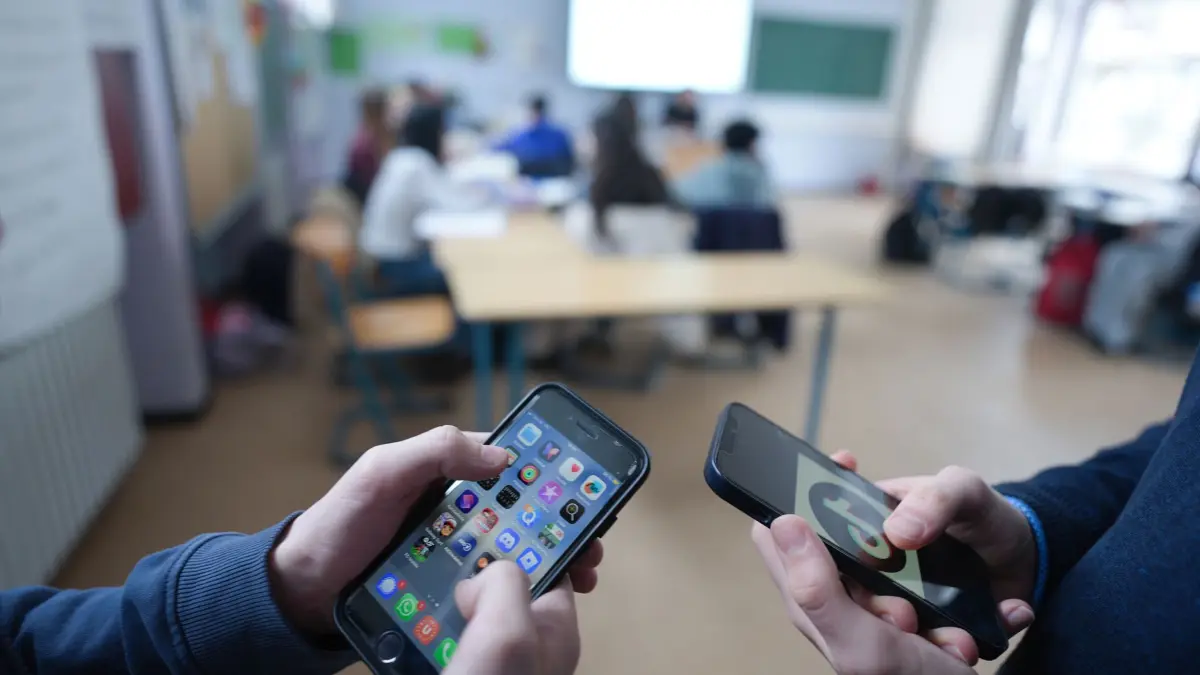 Smartphones in der Schule: ILLUSTRATION - 13.02.2026, Hamburg: Zwei Schüler stehen in einem Klassenraum im Gymnasium und schauen auf ihre Smartphones, auf einem der Displays ist das Logo der Social-Media-Plattform TikTok zu sehen. (zu dpa: «Medienanstalt: Altersgrenzen allein schützen Kinder nicht») Foto: Marcus Brandt/dpa +++ dpa-Bildfunk +++