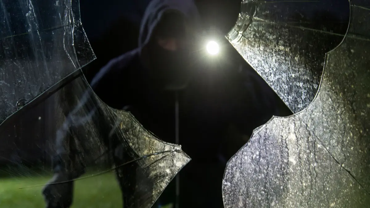 Durch eine eingeschlagene Scheibe blickt ein fiktiver Einbrecher
