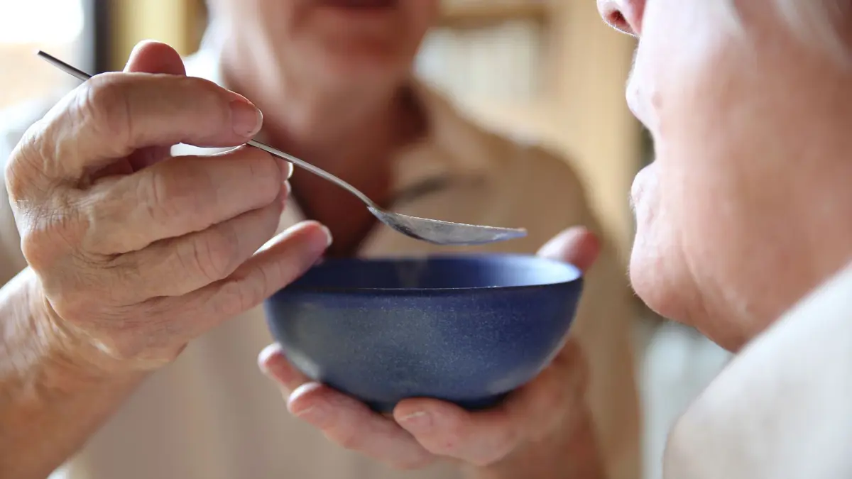 Ein Mann gibt einer zu pflegenden Person etwas zu essen: ILLUSTRATION - Pflegebedürftigkeit kommt längst nicht immer schleichend, sondern kann sich durch Schlaganfall und Co. von jetzt auf gleich einstellen. (zu dpa: «Wie möchte ich im Alter leben? Diese 7 Fragen helfen weiter») Foto: Mascha Brichta/dpa-tmn - Honorarfrei nur für Bezieher des dpa-Themendienstes +++ dpa-Themendienst +++